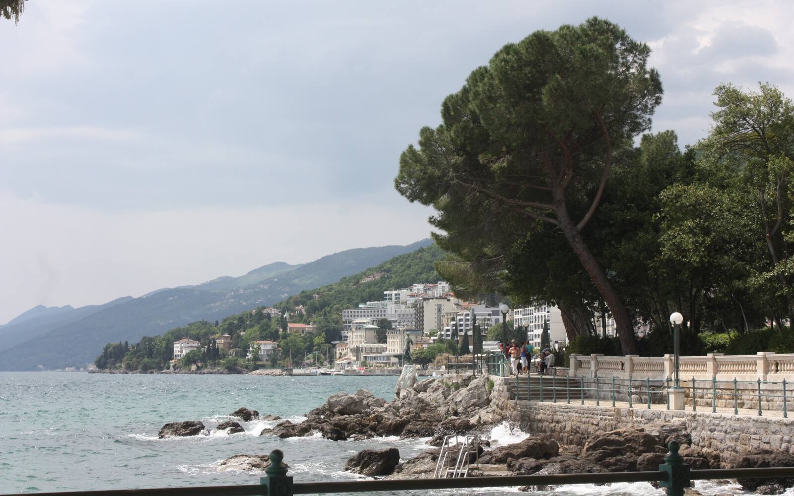Het kustpad Lungomare in Opatija met een witte balustrade, rotsige kust en hotels tegen de groene heuvels